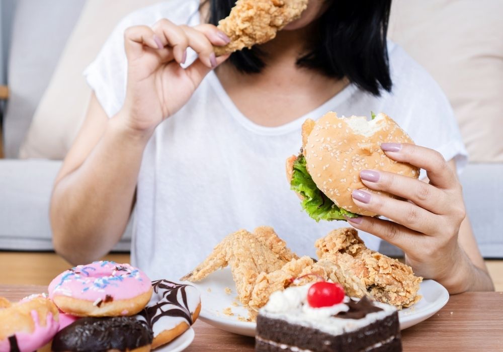 Frau isst Burger, Donuts und Fast Food – stark verarbeitete Lebensmittel können die Darmflora aus dem Gleichgewicht bringen.