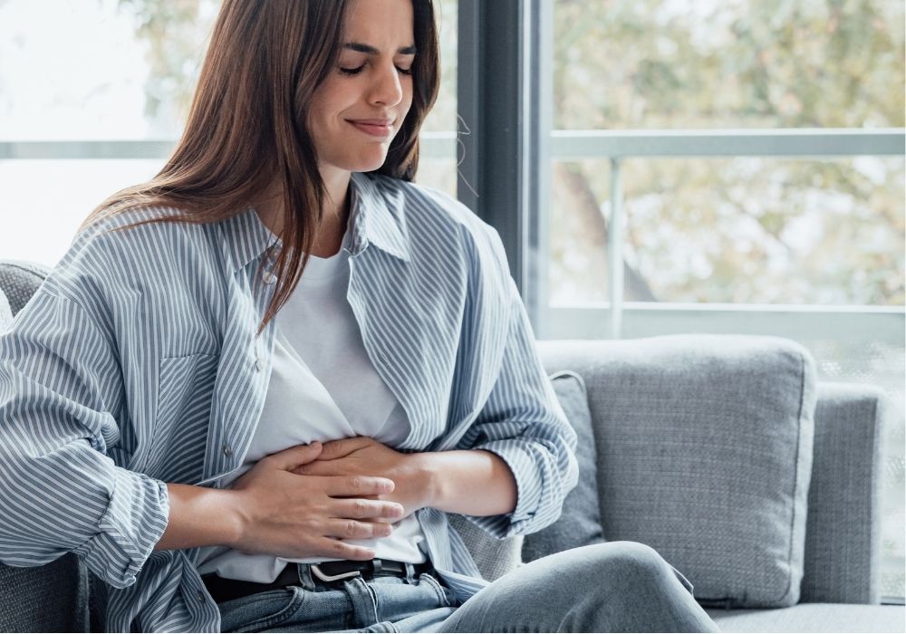 Frau sitzt auf dem Sofa und hält sich den Bauch – mögliche Anzeichen für Verdauungsprobleme oder ein Ungleichgewicht der Darmflora.